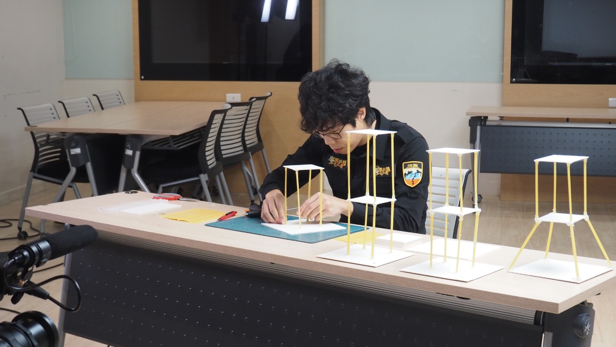 主持人胡子正在用義大利麵條實驗搭起「軟腳蝦建築」。圖／《鹹魚小隊》